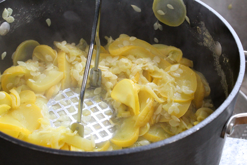 Cooked sliced summer squash in a pot with a potato masher