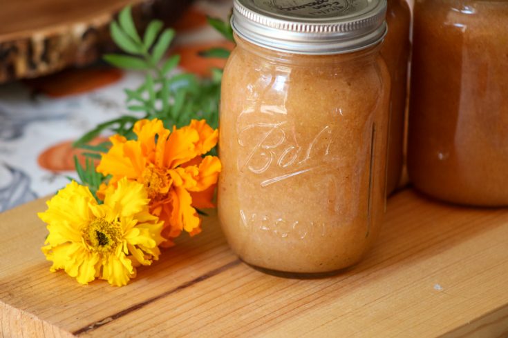 three jars of homemade applesauce made with a kitchenaid mixer in mason jars sitting on a wood board. There are two marigold flowers to the left of the jars.