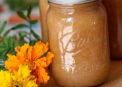 3 quart size mason jars filled with homemade applesauce sitting on a wood board with an orange and yellow marigold flower laying down side them. The jars are on the right with one front and center. The flowers are laying down on the board facing the camera.