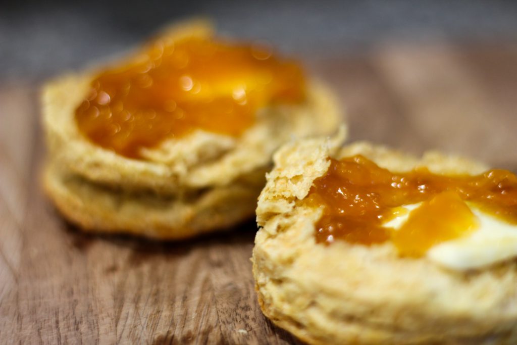 two halves of a biscuit on a wooden cutting board. The biscuits are topped with a slab of butter and homemade peach jam